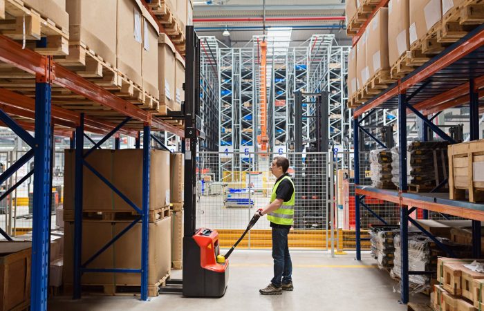 Storehouse man employee in uniform using forklift with box in modern automatic warehouse. Boxes are on the shelves of the warehouse. Warehousing, machinery concept. Logistics in stock.