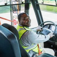 A mature African-American man working as a school bus driver, sitting in the driver's seat wearing his seat belt and a reflective vest, looking over his shoulder at the camera. The bus stop sign is visible in the side view mirror.